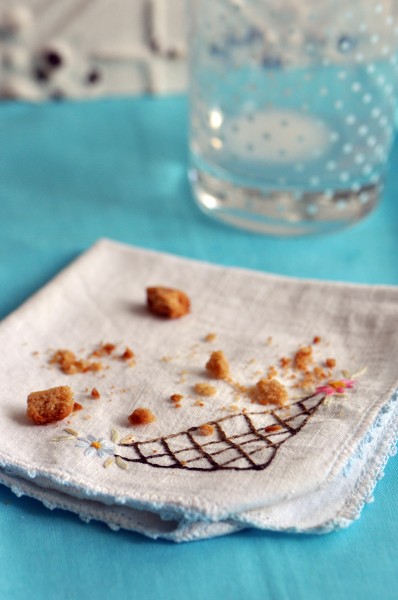 A few peanut butter cookie crumbs sitting on small white embroidered napkin and an empty milk glass in distance