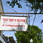 "Sign hanging from pole over top of a shop in red letters says That Shop In Milford"