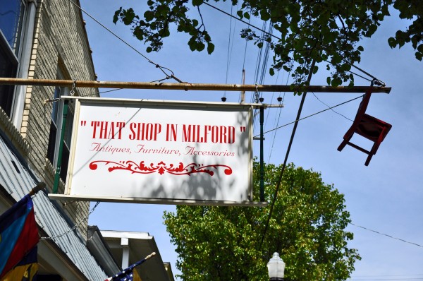 "Sign hanging from pole over top of a shop in red letters says That Shop In Milford"